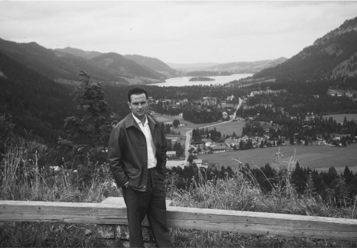 Schwarzweißfotografie eines Mannes, der auf einer Anhöhe steht. Im Hintergrund sieht man ein alpenländisches Panorama mit bewaldeten Hügeln, die einen See umgeben, und einigen Dörfern. Der Mann trägt eine braune Hose, ein weißes Hemd und eine braune Lederjacke. Er hat die Hände in den Hosentaschen und blickt direkt in die Kamera. Es handelt sich bei dem Mann um Arie Katzenstein.