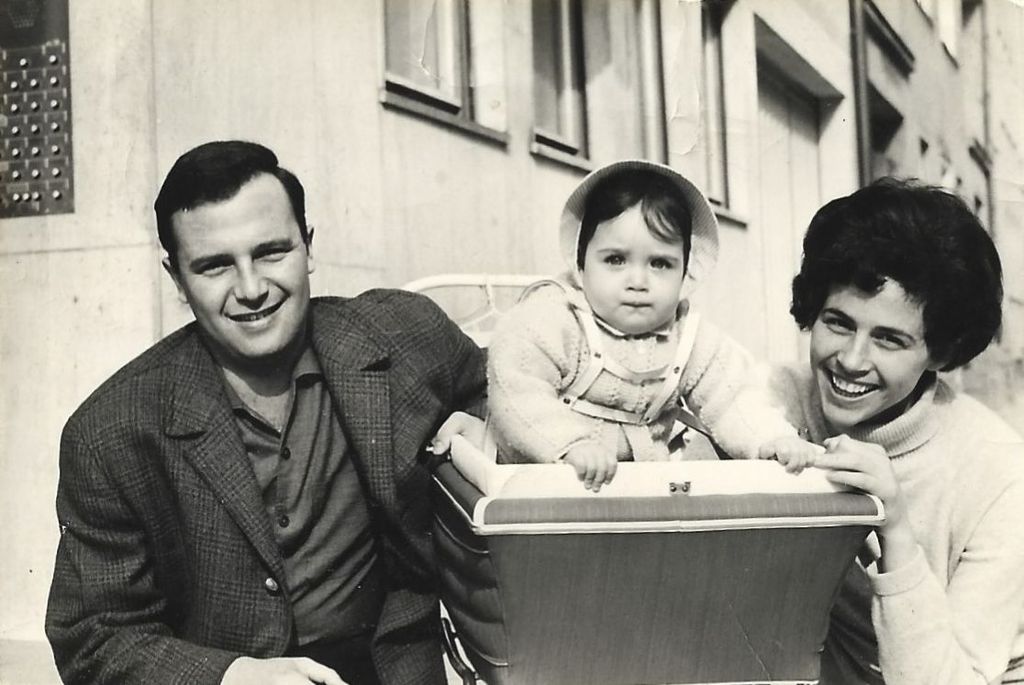 Schwarzweißfotografie einer Familie. In der Mitte steht ein Kinderwagen, aus dem ein etwa einjähriges Kind herausschaut. Auf der einen Seite des Kinderwagens kniet der Vater Arie Katzenstein, er trägt ein Hemd und einen karierten Sakko. Auf der anderen Seite kniet die Mutter Bilha Katzenstein, sie trägt einen hellen Rollkragenpullover. Die Eltern lächeln in die Kamera.