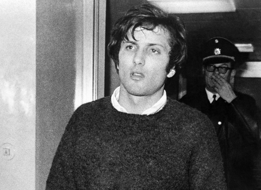 Black and white photograph of a young man walking through a door. He has short dark hair and is wearing a dark woollen jumper with a white shirt. He has a serious look on his face. A police officer can be seen in the background, identified as such by his uniform - peaked cap and coat.