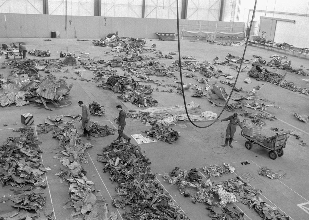 Schwarzweißfotografie mit einer von oben aufgenommenen Ansicht in eine Fabrikhalle. Auf dem Boden der Fabrikhalle sind eine große Anzahl an Flugzeugtrümmern in vorgegebenen Markierungen ausgelegt. Zwischen den Trümmerteilen bewegen sich drei Männer in Arbeitsanzügen.