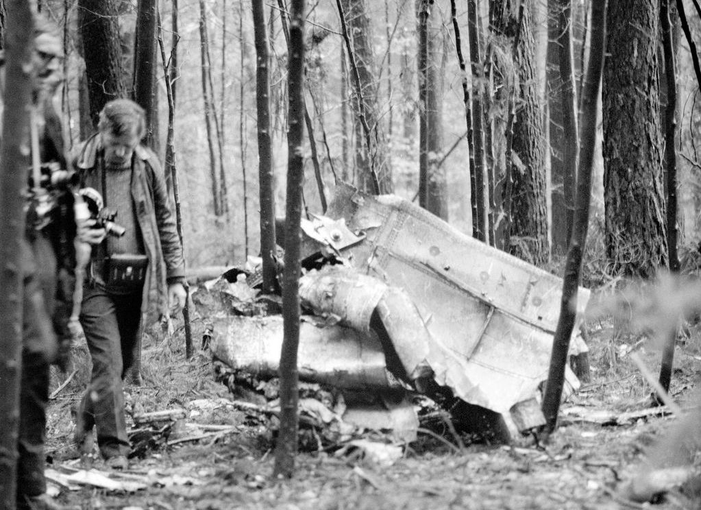 Schwarzweißfotografie einer Flugzeug-Absturzstelle im Wald. Zwischen den Bäumen liegen Trümmerteile auf dem Waldboden. Neben den Trümmern sind zwei Männer mit Fotoapparaten zu sehen.