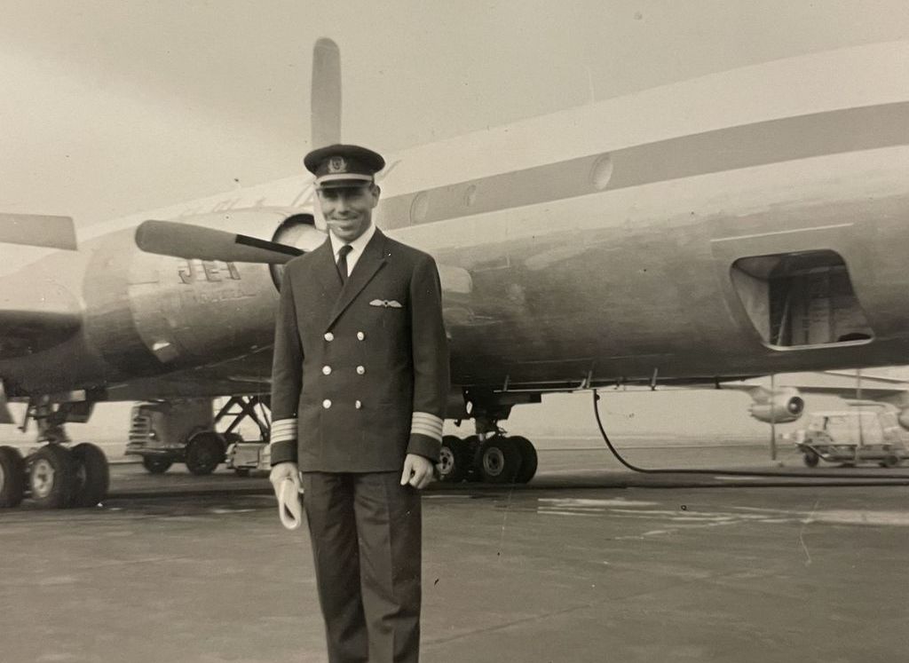 Sepiafarbene Fotografie des Piloten Uriel Cohen auf dem Rollfeld eines Flughafens. Der Mann trägt Uniform, bestehend aus Kapitänsmütze, einem Anzug mit Abzeichen an Brust und Armen, Hemd und Krawatte. Er blickt lächeln in die Kamera. Hinter ihm ist ein auf dem Rollfeld geparktes Flugzeug zu sehen, das gerade betankt wird.