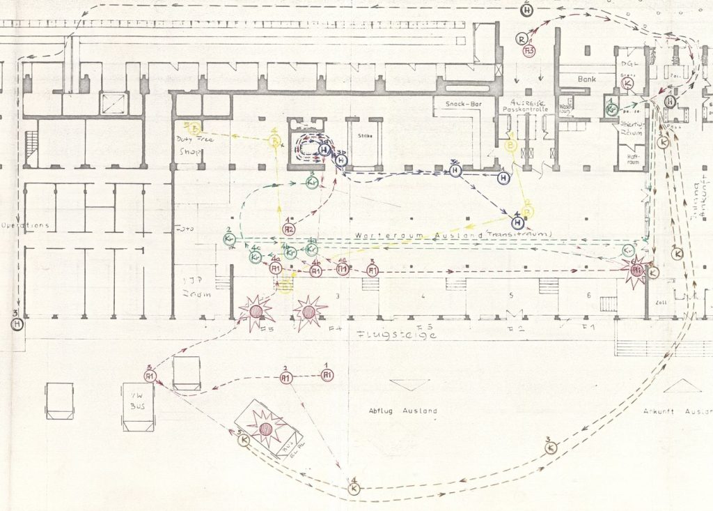 Architektonische Planskizze eines Gebäudegrundrisses. Die Linien sind mit grauem Stift auf beigem Papier ausgeführt. Verschiedene Gebäudeteile sind beschriftet. Aus den Beschriftungen lässt sich erkennen, dass es sich bei dem Bau um Teile eines Flughafengebäudes handelt. Im Zentrum der Zeichnung liegt der Warteraum für Auslandsflüge. In den Plan sind gestrichelte Linien in unterschiedlichen Farben eingezeichnet, die von Pfeilen und Kreisen unterbrochen sind und die Laufwege darstellen sollen. Die Kreise sind mit Nummern und Zahlen beschriftet. Zudem sind vier sternförmige Markierungen auf der Karte eingezeichnet.