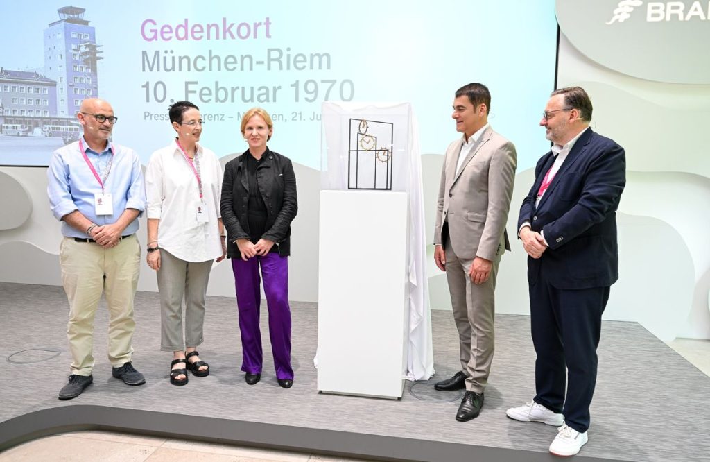 Foto der Pressekonferenz zur Vorstellung des Gedenkorts München-Riem 10. Februar 1970. Um ein Modell des Erinnerungsortes haben sich auf einer Bühne fünf Personen gruppiert. Es handelt sich von links gelesen um: Ofer Katzenstein und Miki Dror, Künstlerin Alicja Kwade, Brainlab CEO Stefan Vilsmeier und Kulturreferent Anton Biebl.