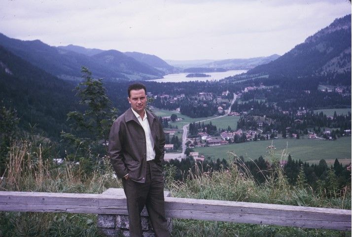 Photograph of a man standing on a hill. In the background is an Alpine panorama with wooded hills surrounding a lake and some villages. The man is wearing brown trousers, a white shirt and a brown leather jacket. He has his hands in his trouser pockets and is looking directly at the camera. The man is Arie Katzenstein.