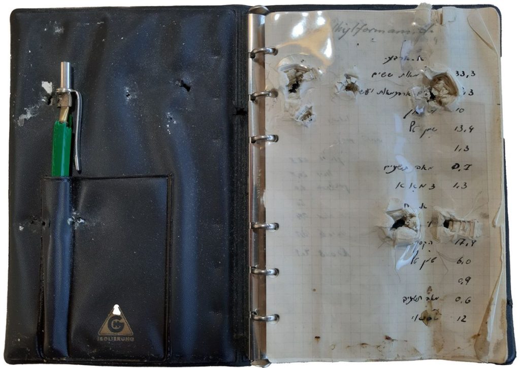 Image of an open leather notebook. A green ballpoint pen is stuck in the cover. The pages of the notebook are torn and have holes in several places.