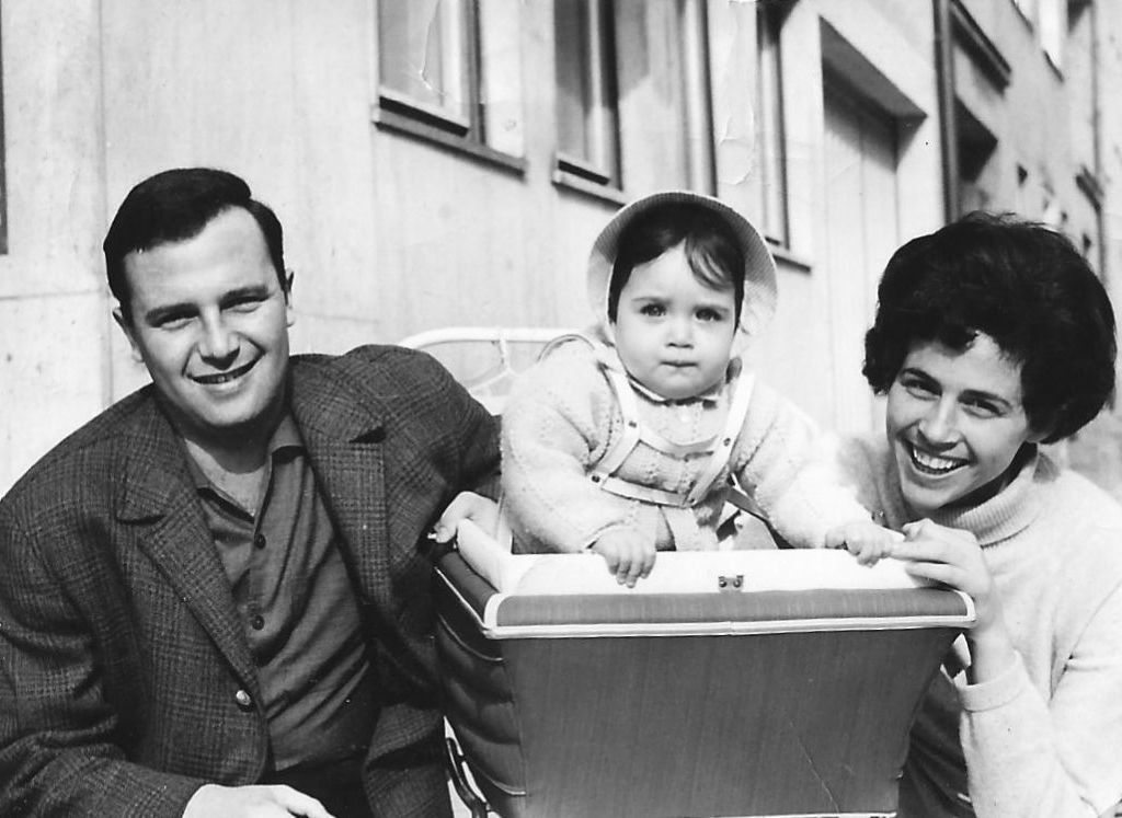 Black and white photograph of a family. In the middle is a baby carriage with a roughly one-year-old child looking out of it. On one side of the baby carriage kneels the father, Arie Katzenstein, wearing a shirt and a checked jacket. On the other side kneels the mother, Bilha Katzenstein, wearing a light-colored turtleneck sweater. The parents smile at the camera.