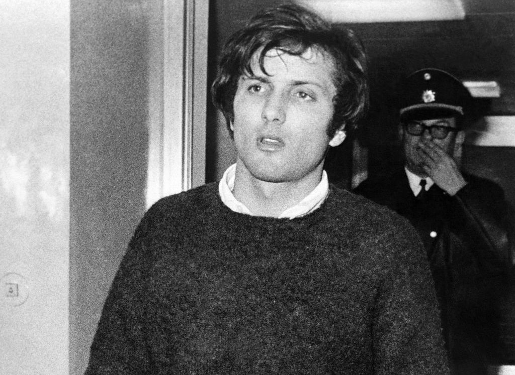 Black and white photograph of a young man walking through a door. He has short dark hair and is wearing a dark woollen jumper with a white shirt. He has a serious expression on his face. In the background is a police officer, recognisable by his uniform - peaked cap and coat.