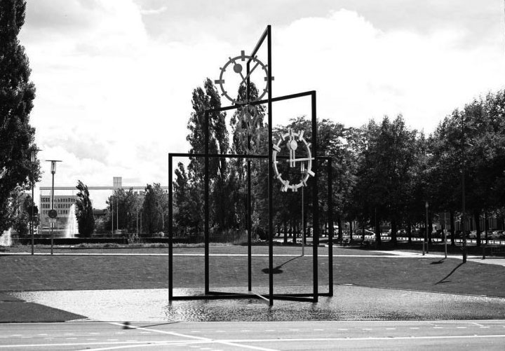 Black and white photograph of a work of art that is set up outdoors in a public space. The artwork by the artist Alicja Kwade consists of three steel rectangles of different heights, which are mounted inside each other at different angles. Between the steel beams are three clock faces with hands indicating different times. The steel construction stands in a shallow pool of water. Footpaths, trees and other buildings can be seen in the background.