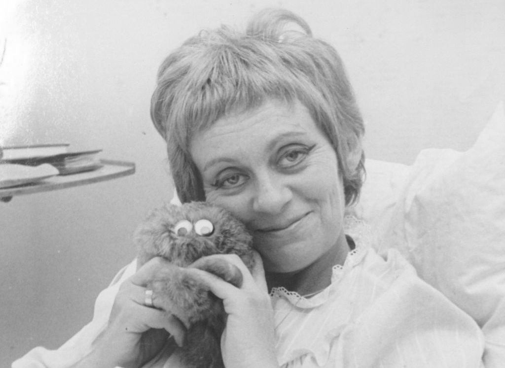 Black and white photograph taken in a hospital room. The picture shows a close-up of a woman with short hair in her 40s, dressed in a white nightgown with a lace collar. She is the actress Hanna Maron. She is sitting in a hospital bed with her upper body propped up on pillows, holding a cuddly toy in both hands and pressing it against her cheek. A little exhausted, she smiles at the camera. The pull-out tray of a bedside table can be seen in the background.