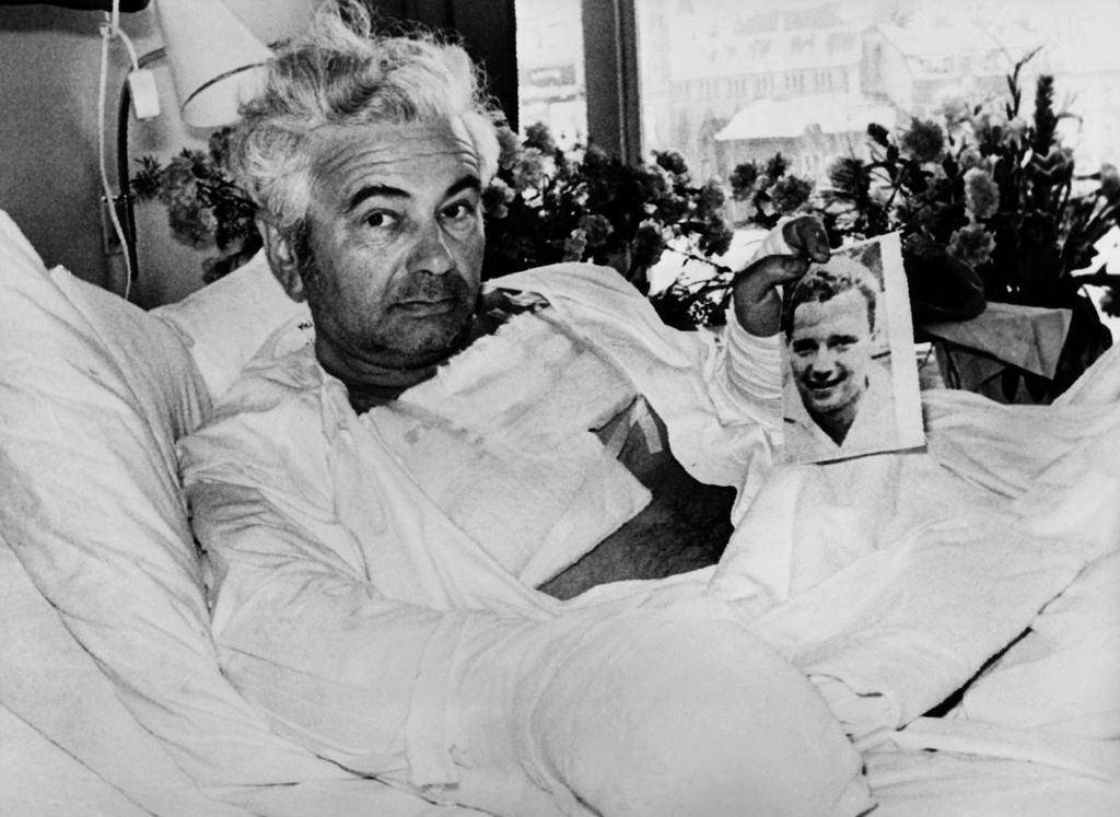 Black and white photograph showing a man with grey hair in a hospital room. The man is sitting in a hospital bed with his upper body slightly raised on a pillow and turned towards the camera; he is covered up to his stomach with a white blanket. He is bandaged on several parts of his body, such as his chest and arms. He holds a photograph of a smiling young man in front of the camera. A large window can be seen in the background, through which a snow-covered street view is visible. On the windowsill are a row of bouquets of flowers.