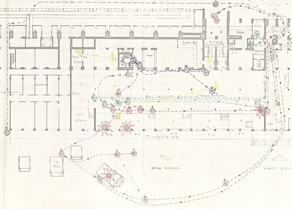 Architectural sketch of a ground plan of a building. The lines are drawn in grey pencil on beige paper. Various parts of the building are labelled. From the inscriptions it can be deduced that the building is part of an airport building. The waiting room for international flights is in the centre of the drawing. The plan shows dashed lines in different colours, interrupted by arrows and circles, which are intended to represent walkways. The circles are labelled with numbers and figures. There are also four star-shaped markings on the map.