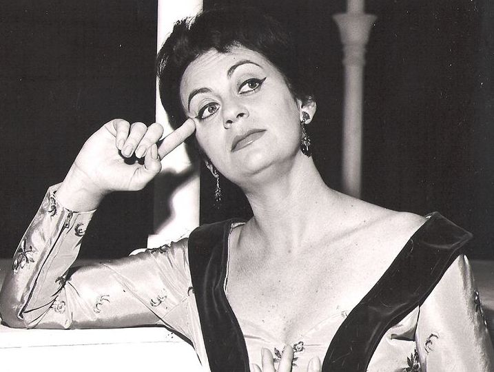 Black and white photograph of a woman. The woman has short dark hair, her eyes are heavily accentuated with black kohl and she is wearing a dress with a low neckline. She is the actress Hanna Maron. She is resting her head on one hand and holding her heart with the other.