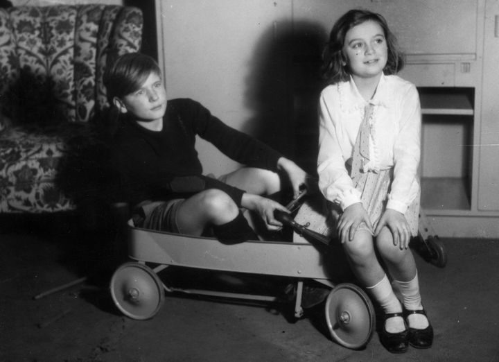 Black and white picture with a boy and a girl in the centre. The picture was taken in an interior, probably a living room. In the background on the wall are a floral armchair and a cupboard. Both children are sitting on a metal, steerable handcart. The boy is sitting in the cart, his hair is parted to the side, he is dressed in a black jumper, shorts and knee-high socks. The girl has shoulder-length hair, also parted and pinned to one side with a hair clip. She is wearing a white blouse with a narrow long scarf, a pleated skirt, knee-high socks and black patent leather shoes. She is sitting at the front of the carriage, her hands resting on her knees. The children's gaze is directed sideways towards an object or person outside the frame.