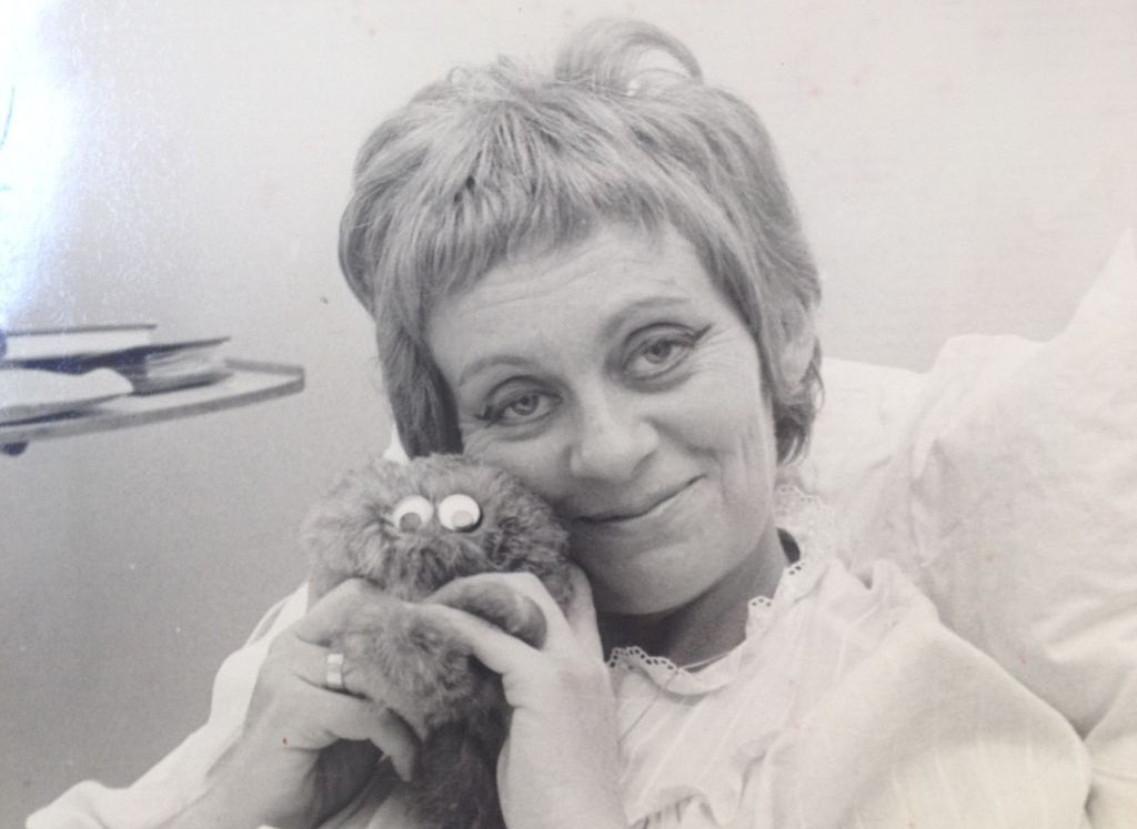 Black and white photograph taken in a hospital room. The picture shows a close-up of a woman with short hair in her 40s, dressed in a white nightgown with a lace collar. She is the actress Hanna Maron. She is sitting in a hospital bed with her upper body propped up on pillows, holding a cuddly toy in both hands and pressing it against her cheek. A little exhausted, she smiles at the camera. The pull-out tray of a bedside table can be seen in the background.