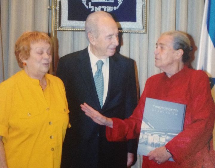 Photograph of a group of three people, one man and two women, at an official event. The group is standing in front of a curtain, the national emblem of the State of Israel can be seen in the background. The man standing in the centre is former Israeli President Shimon Peres. On one side he is flanked by a lady with short hair and in a yellow dress, the actress Hanna Maron. On the other side is a lady in a red suit holding a book about the Cameri Theatre.