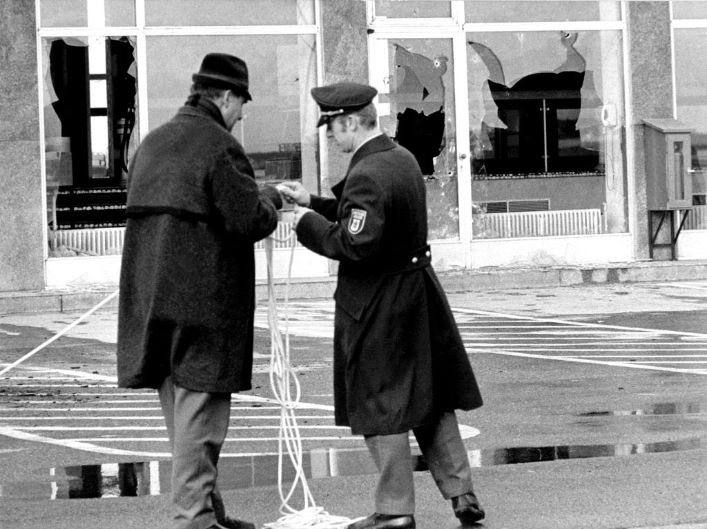 Black and white photograph showing two men in the foreground, seen from behind, standing on a kind of tarmac with white floor markings. The ground floor of a building can be seen in the background. The large window fronts are shattered and have large holes through which one can see into the hall-like interior of the building. The two men are wearing black coats and hats. One of the men is identified as a Munich police officer by a shoulder badge on his coat and his peaked cap. They are holding a white rope in their hands, which they appear to be trying to untangle.