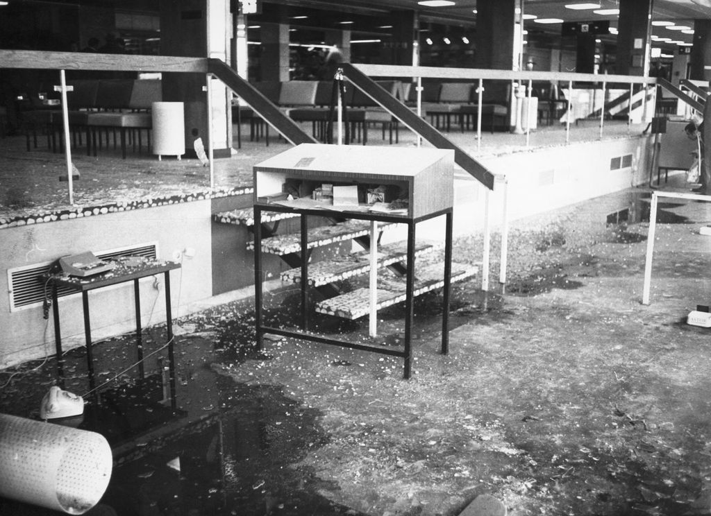 Black and white photograph showing the view into the destroyed waiting hall of the former Munich-Riem Airport. The hall has two levels connected by a few steps, with a wooden railing separating the sections: In the lower area there are a few objects, such as a small table and a vitrine, in the upper waiting area there are rows of upholstered chairs. The hall shows signs of severe devastation. There is a large amount of broken glass on the floor and tables, and objects such as a telephone and rubbish bin have been thrown onto the floor. Parts of the floor are also covered by water.