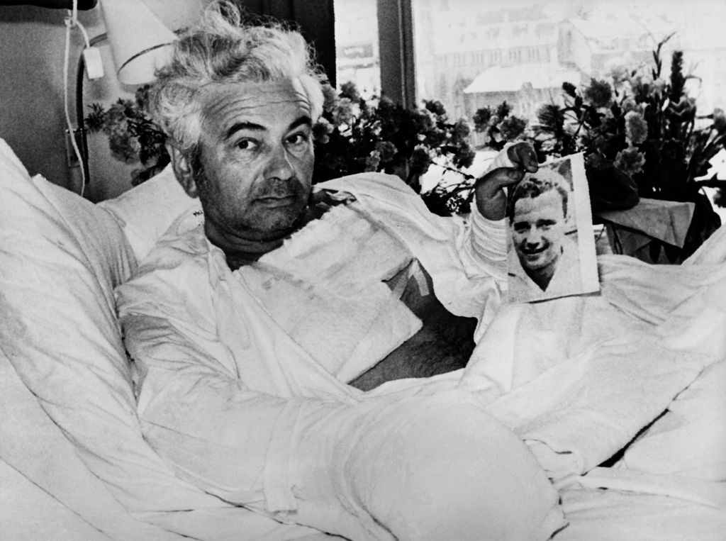 Black and white photograph showing a man with grey hair in a hospital room. The man is sitting in a hospital bed with his upper body slightly raised on a pillow and turned towards the camera; he is covered up to his stomach with a white blanket. He is bandaged on several parts of his body, such as his chest and arms. He holds a photograph of a smiling young man in front of the camera. A large window can be seen in the background, through which a snow-covered street view is visible. On the windowsill are a row of bouquets of flowers.