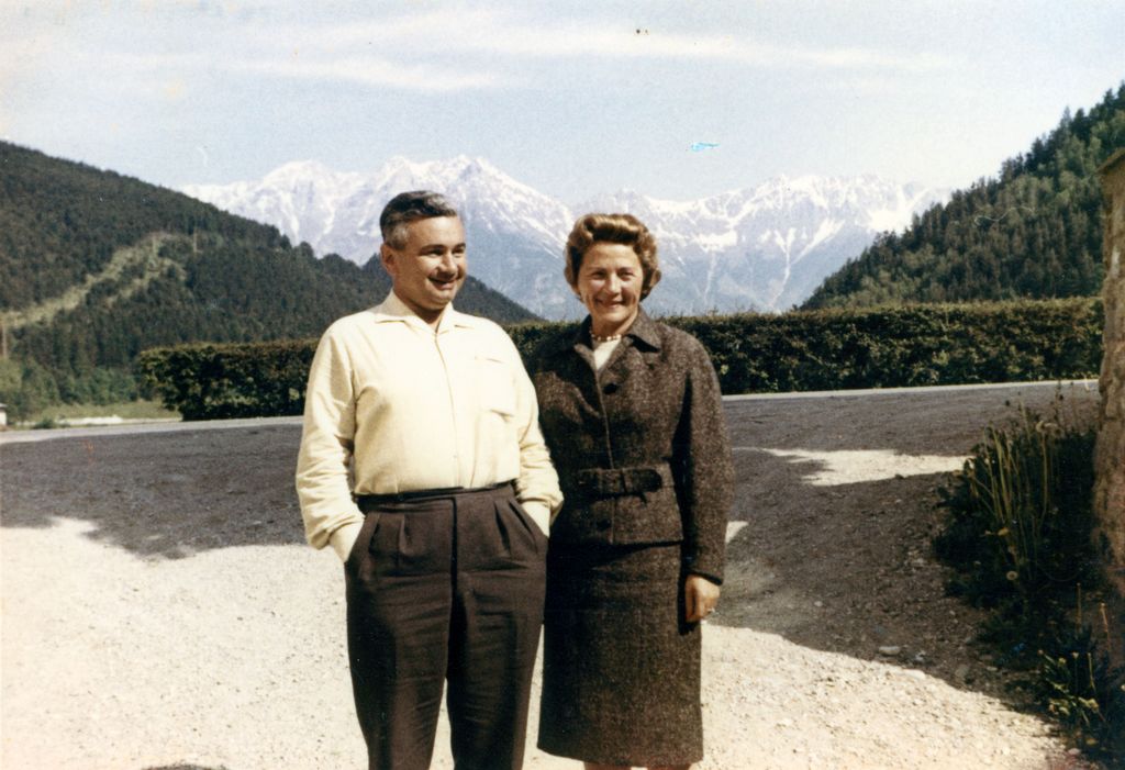 Colour photograph of an elderly couple in front of a panorama of the Alps. The man, with grey hair, is wearing brown trousers and a white shirt, while the woman is wearing a brown woollen suit and has wavy hair. They are standing on a path in the countryside, with wooded hills and a snow-capped mountain range in the background.