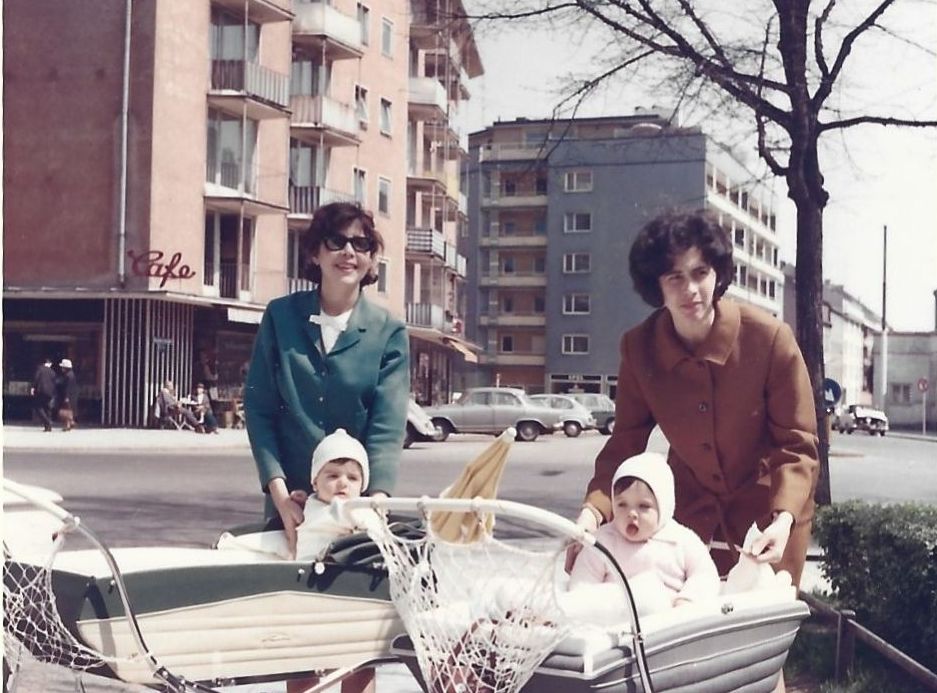 Photograph of two women with prams in a public square. The two toddlers are sitting upright in their prams. A woman in a brown costume is leaning slightly over the pram; she is Bilha Katzenstein. Multi-storey houses and a few parked cars can be seen in the background.