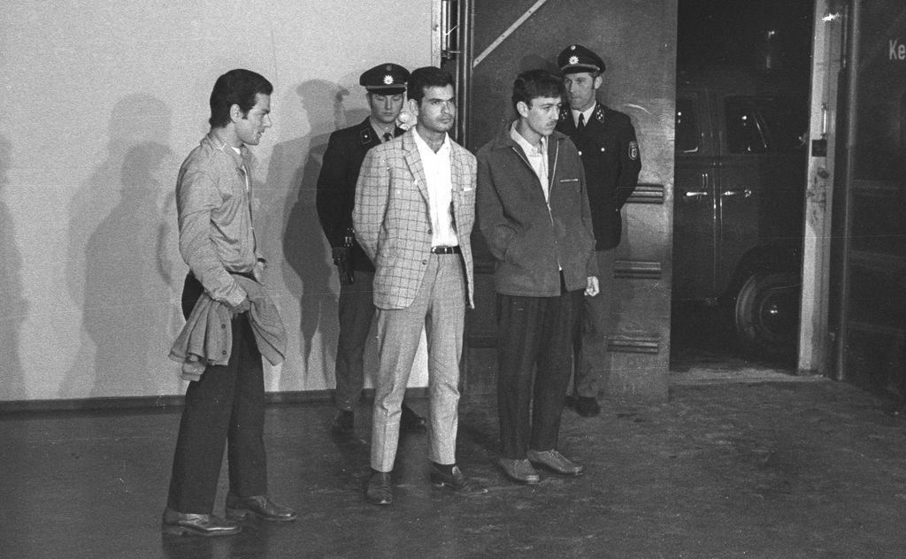 Black and white photograph of several people, taken in a hall-like interior. Three young men of Arab origin are standing next to each other with their hands in their pockets or crossed behind their backs. Behind them are two uniformed police officers.