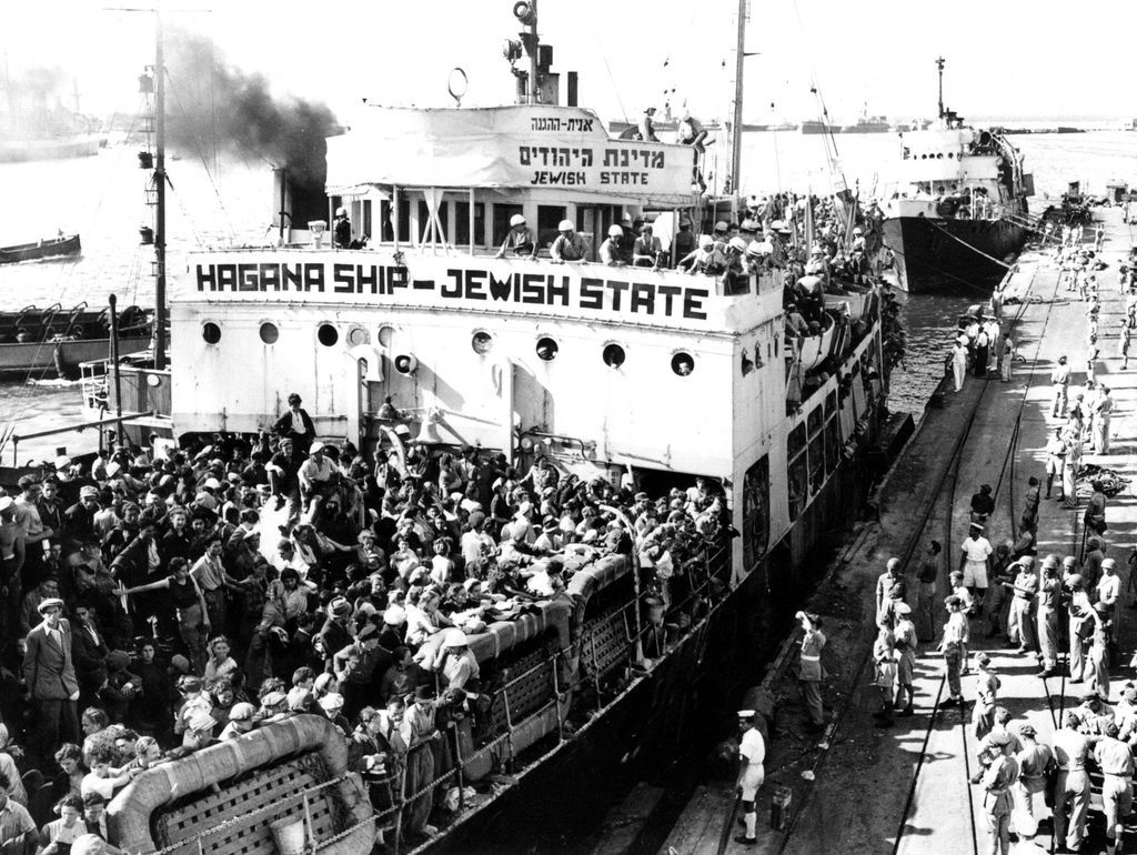 Black and white photograph of a ship crowded with people docked in a harbour basin. The words 'Hagana Ship - Jewish State' are written on the ship. A large number of people are crowded together on the deck of the ship. A number of uniformed people are standing in the harbour looking at the ship.