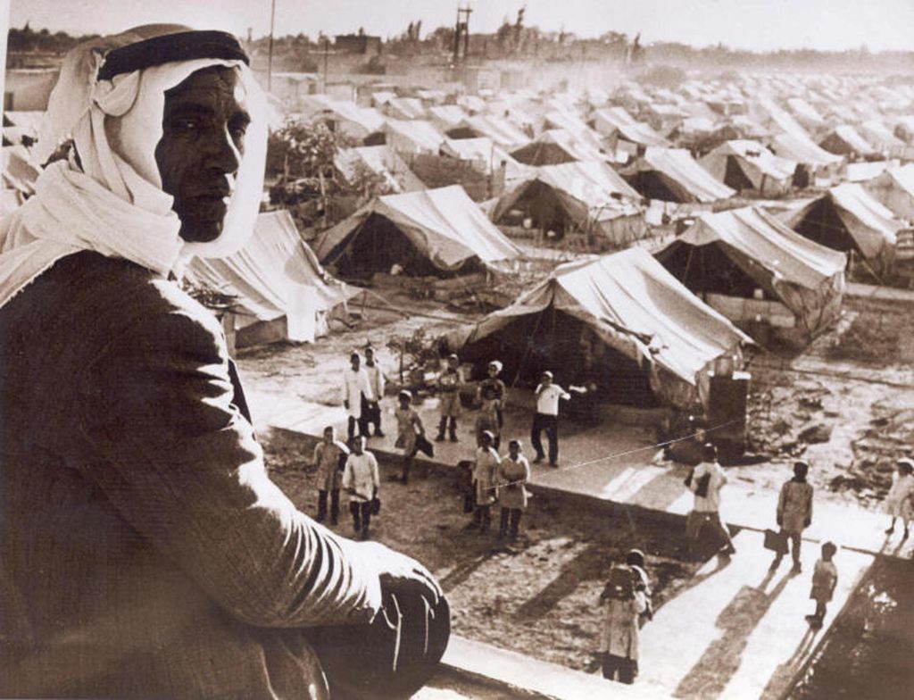 Sepia-toned photograph of a refugee camp, taken from a hilltop. In the foreground of the photograph, a man wearing a kufiya headdress sits on the hill looking towards the camera. Many rows of tents can be seen in the valley, with paths running between them. At the foot of the hill are children looking up at the camera.