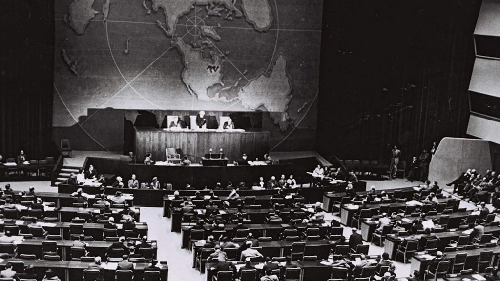 Black and white photograph of the UN General Assembly in New York, 1947, taken from above. The rows of seats in the General Assembly are seen from behind.