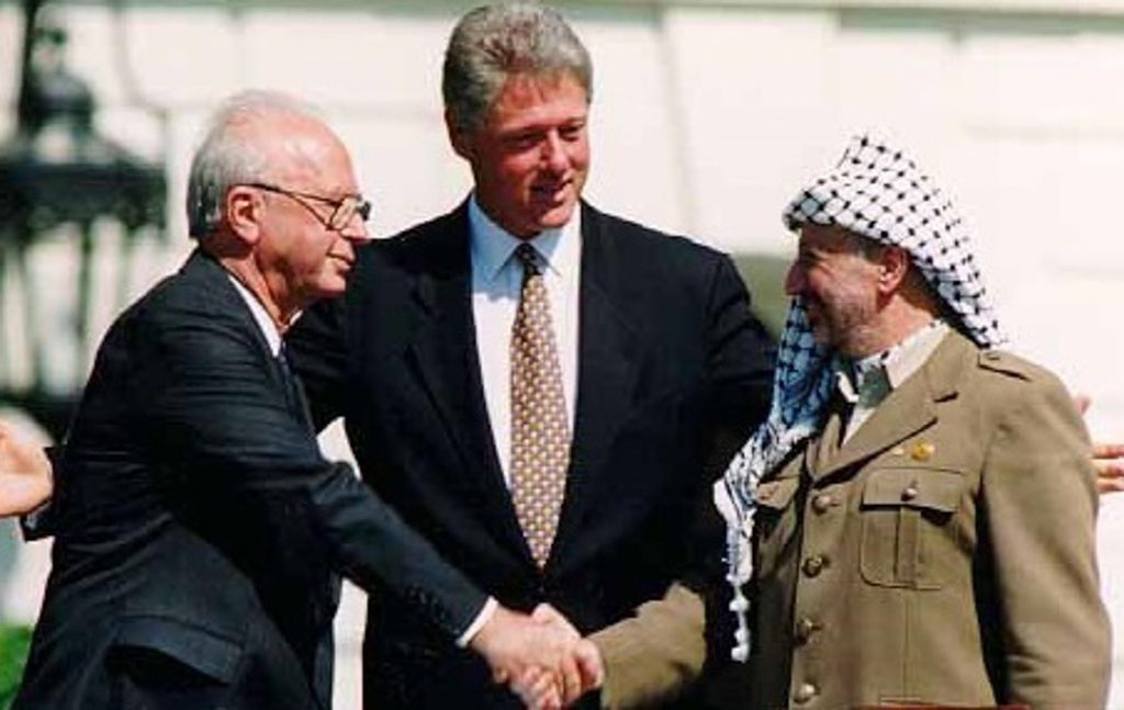 Photograph taken at the signing of the ‘Oslo I’ accord on 13.9.199 . Two men are facing each other and shaking hands. They are the Prime Minister of Israel, Yitzchak Rabin, and the Chairman of the PLO, Yasser Arafat. Behind them stands another man, both arms slightly outstretched in a unifying gesture. It is US President Bill Clinton.