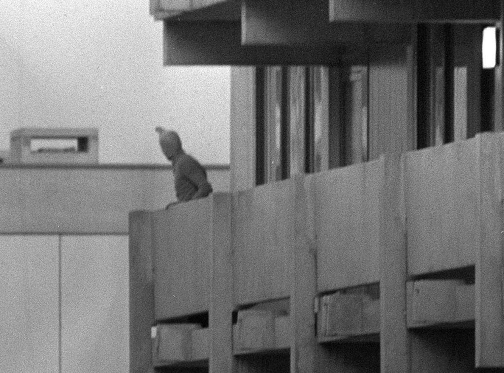 Black and white photograph taken during the attack on the 1972 Olympics. The picture shows part of a building. The building is surrounded by a concrete balcony. A man wearing a stocking mask is standing on the balcony, looking over the railing.