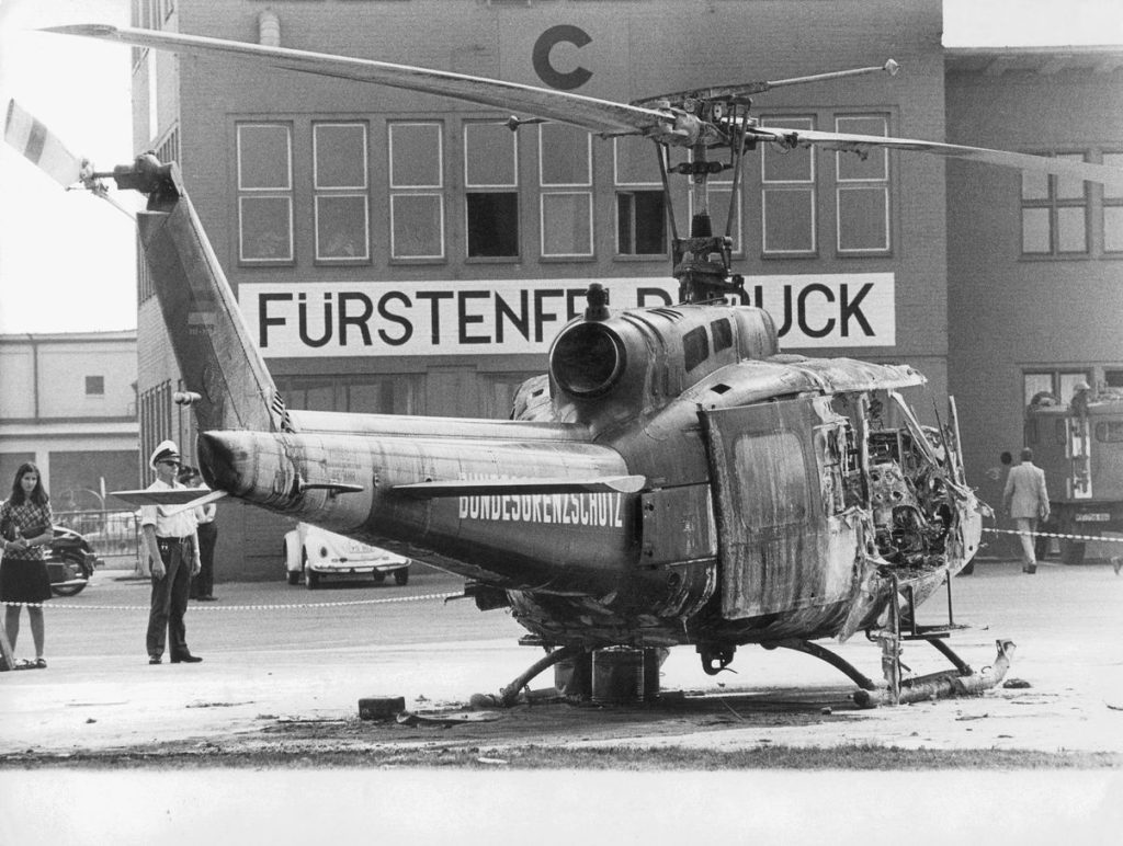 Black and white photograph of a burnt out helicopter on an airfield. Bundesgrenzschutz" is written on the back of the helicopter. The area around the helicopter is cordoned off with tape, some people are standing around the helicopter. Airfield buildings can be seen in the background, one building is labelled with the name of the airfield 'Fürstenfeldbruck'.