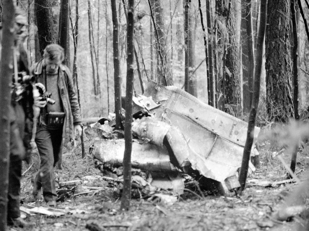 Black and white photograph of an airplane crash site in the forest. Debris is lying on the forest floor between the trees. Two men with cameras stand beside the debris.