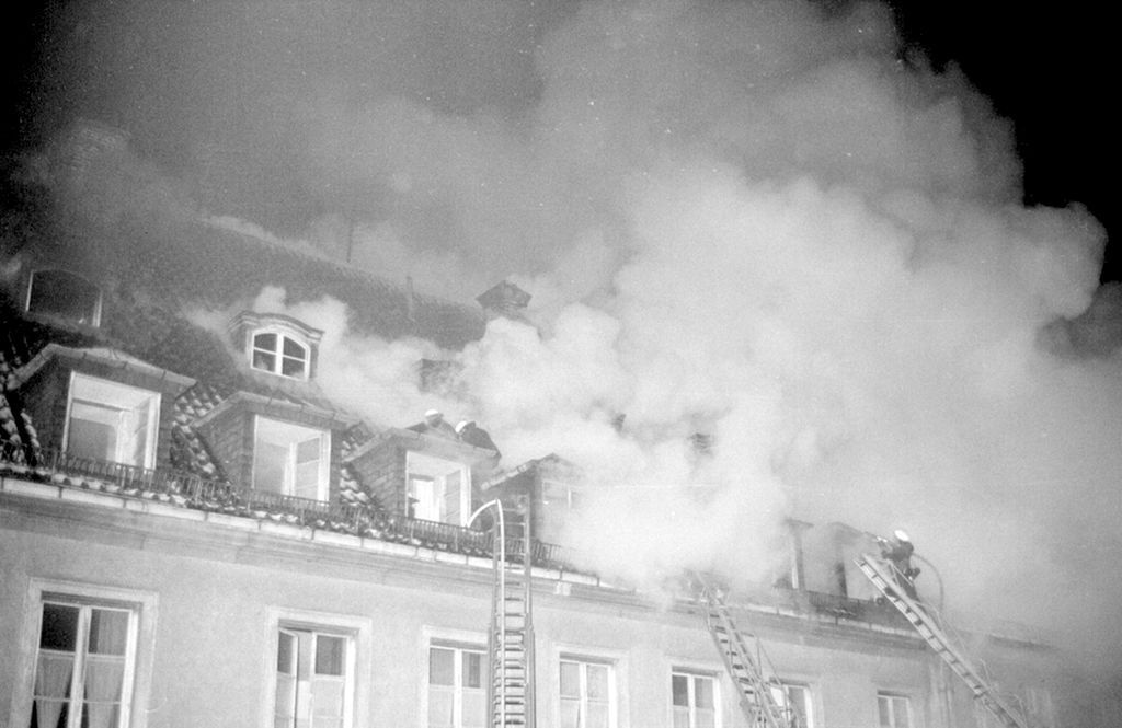 Black and white photograph of a burning roof being extinguished by the fire brigade. Smoke rises from the skylights and the roof. Three fire ladders are leaning against the roof, three firemen are on the roof extinguishing the fire.