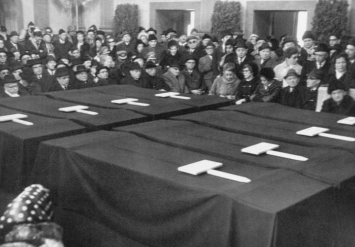 Black and white photograph of a funeral service being held in an interior room. In the center of the picture, you can see eight coffins placed next to each other in two rows of four. The coffins are covered with black cloths. Many people are sitting and standing around the coffins in several rows one behind the other. The mourners are wearing dark clothing, the men are wearing head coverings.