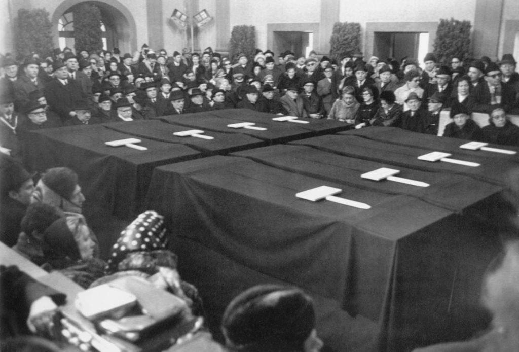 Black and white photograph of a funeral service being held in an interior room. In the center of the picture, you can see eight coffins placed next to each other in two rows of four. The coffins are covered with black cloths. Many people are sitting and standing around the coffins in several rows one behind the other. The mourners are wearing dark clothing, the men are wearing head coverings, as are many of the women.