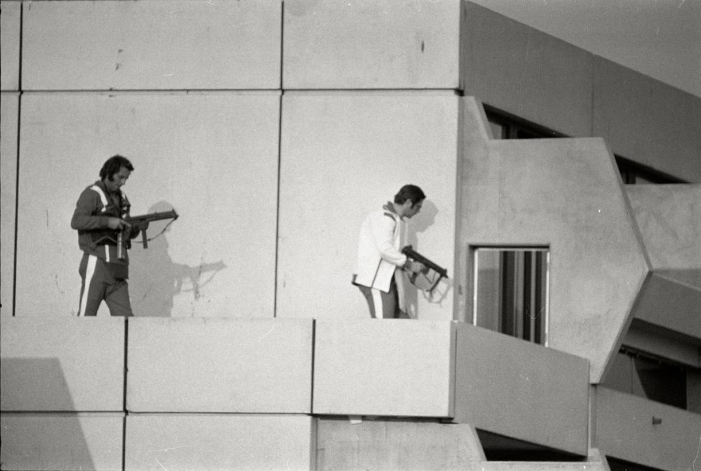 Black and white photograph taken during the attack on the 1972 Olympics. The picture shows part of a concrete building with a surrounding balcony. On the balcony are two men in tracksuits holding submachine guns.