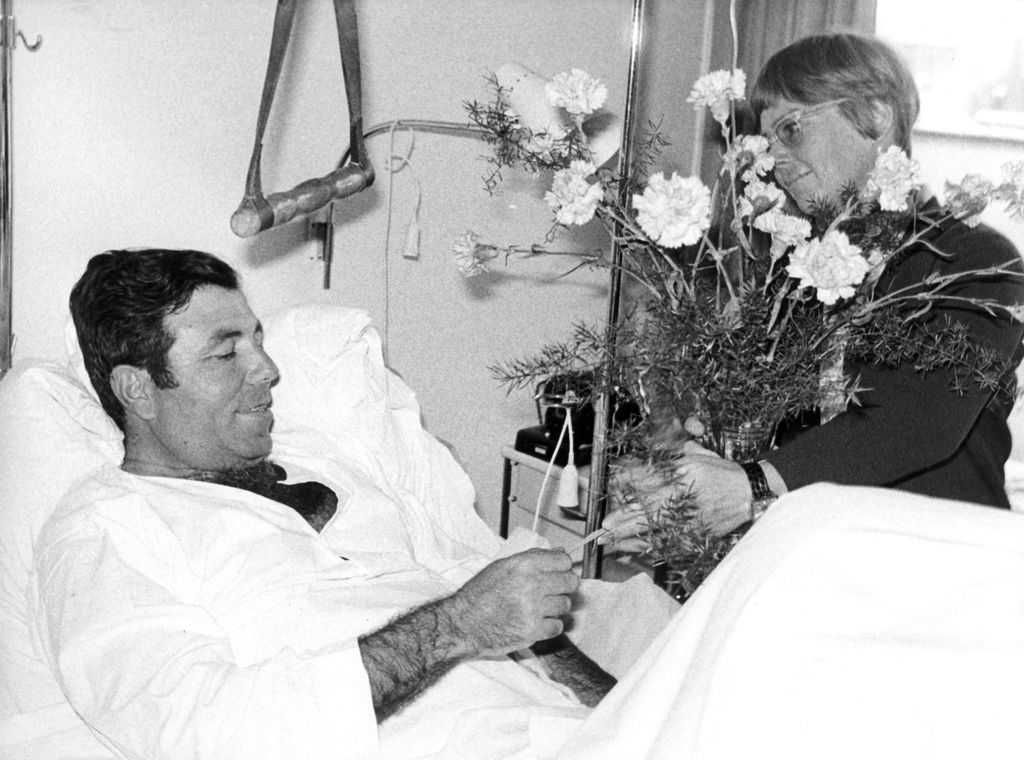 Black and white photograph of a man in a hospital room. He is sitting lightly propped up on a pillow in a hospital bed, wearing a white nightdress and covered up to his stomach with a blanket. Next to the bed is a woman with short hair and glasses, holding a large bouquet of flowers. They are Uriel Cohen and his wife Inga.