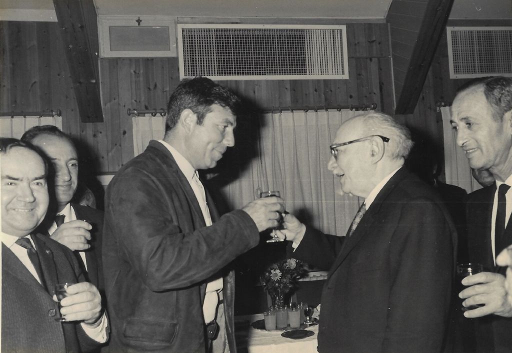 Black and white photograph of a formal event. At the centre of the photograph are two men facing each other, clinking glasses. They are Uriel Cohen and the President of Israel, Salman Shasar. They are surrounded by three men who are also holding glasses.