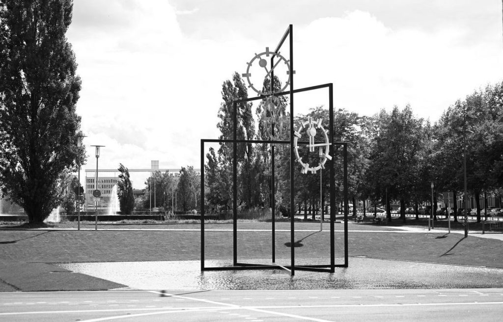 Black and white photograph of a work of art that is set up outdoors in a public space. The artwork by the artist Alicja Kwade consists of three steel rectangles of different heights, which are mounted inside each other at different angles. Between the steel beams are three clock faces with hands indicating different times. The steel construction stands in a shallow water basin. Footpaths, trees and other buildings can be seen in the background.