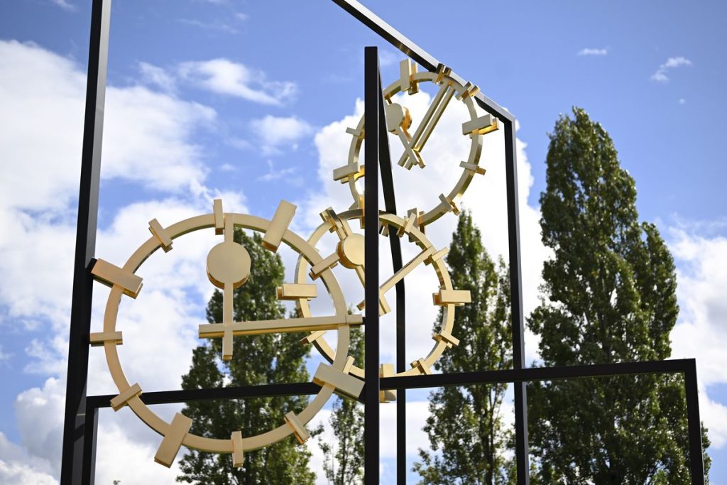 Close-up photograph of a work of art set up outdoors in a public space. The artwork by the artist Alicja Kwade consists of three black steel rectangles of different heights, which are mounted inside each other at different angles. Between the steel beams are three golden clock faces with hands indicating different times. The sky and deciduous trees can be seen in the background.