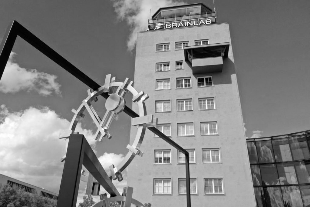 Black-and-white-photograph of a work of art set up outdoors and a multi-storey tower-like building behind it. The artwork by the artist Alicja Kwade consists of black steel rectangles of different heights, which are mounted inside each other at different angles. Between the steel girders are clock faces with hands indicating different times. Behind it is a tower, the former airport tower of Munich-Riem Airport. The company logo “Brainlab” is attached to the tower.