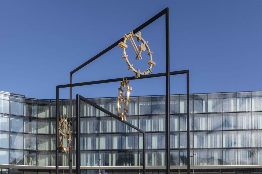 Close-up photograph of a work of art set up outdoors in a public space. The artwork by the artist Alicja Kwade consists of three black steel rectangles of different heights, which are mounted inside each other at different angles. Between the steel beams are three golden clock faces with hands indicating different times. The sky and deciduous trees can be seen in the background.