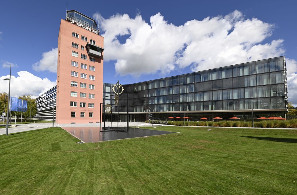 Wide-angle photograph of a large building complex. This consists of a central red tower-like building, the former airport tower of Munich-Riem Airport. Two elongated modern glass office complexes are arranged around the tower. Today the buildings house the headquarters of Branlab AG, the company logo is mounted on the tower. On the forecourt in front of the buildings, a work of art has been erected between the lawns. The artwork by the artist Alicja Kwade consists of black steel beams and three golden clock faces mounted between the steel arms. The artwork stands on a shallow water basin.