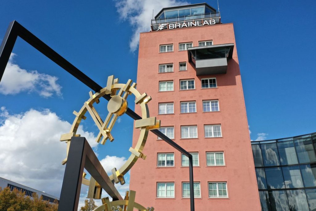 Close-up photograph of a work of art set up outdoors and a multi-storey tower-like building behind it. The artwork by the artist Alicja Kwade consists of black steel rectangles of different heights, which are mounted inside each other at different angles. Between the steel girders are golden clock faces with hands indicating different times. Behind it is a red tower, the former airport tower of Munich-Riem Airport. Today it houses the headquarters of Branlab AG. The company logo “Brainlab” is attached to the tower.