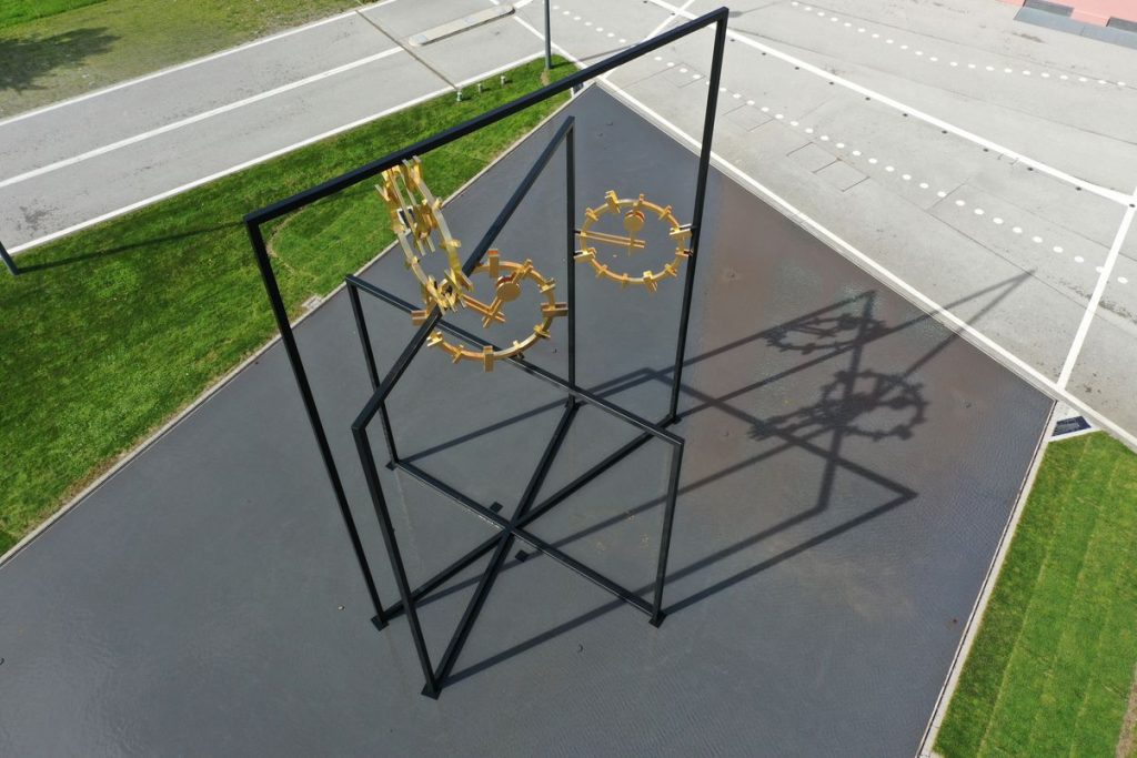 Photograph of a work of art set up outdoors in a public space. The photograph is taken from above from a bird's eye view. The artwork by the artist Alicja Kwade consists of three black steel rectangles of different heights, which are mounted inside each other at different angles. Between the steel beams are three golden clock faces with hands indicating different times. The steel construction stands in a shallow water basin. Around the water basin are walkways with white markings and lawns.