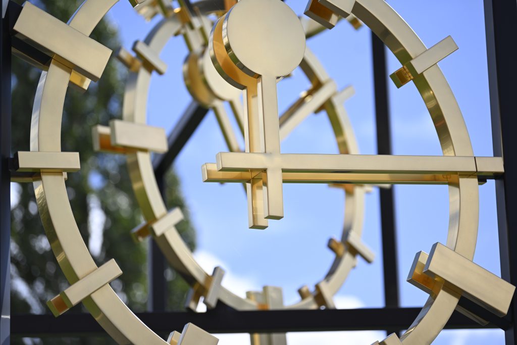 Close-up photograph of a work of art. The artwork by the artist Alicja Kwade consists of a black steel construction in which three gold-plated clock faces are mounted, the hands of which indicate different times. The photograph shows a close-up of the three clock faces appearing one behind the other.