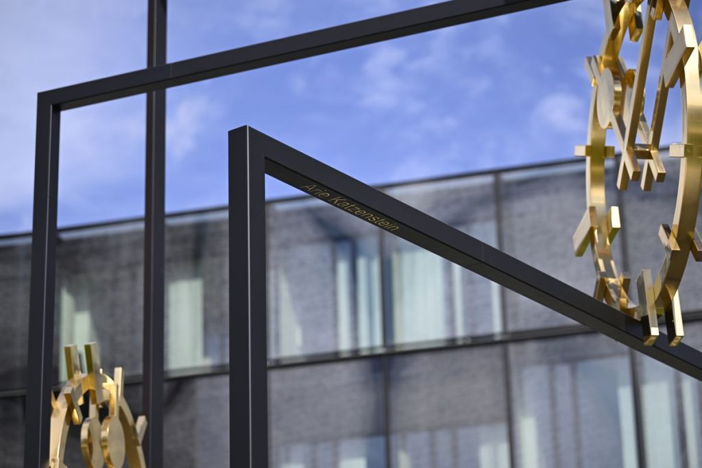 Close-up photograph of a work of art set up outdoors. The artwork by the artist Alicja Kwade consists of a black steel construction in which golden clock faces are mounted. The dials show different times. The name “Arie Katzenstein” is engraved in gold lettering in a steel frame. Tall office buildings with a glass façade can be seen in the background.