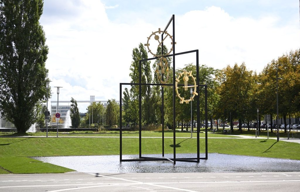 Photograph of a work of art that is set up outdoors in a public space. The artwork by the artist Alicja Kwade consists of three black steel rectangles of different heights, which are mounted inside each other at different angles. Between the steel beams are three golden clock faces with hands indicating different times. The steel construction stands in a shallow water basin. Footpaths, trees and other buildings can be seen in the background.