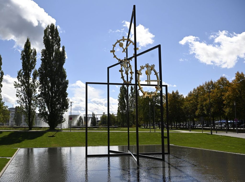 Photograph of a work of art that is set up outdoors in a public space. The artwork consists of three black steel rectangles of different heights, which are mounted inside each other at different angles. Between the steel beams are three golden clock faces with hands indicating different times. The steel construction stands in a shallow water basin. Footpaths, trees and other buildings can be seen in the background.